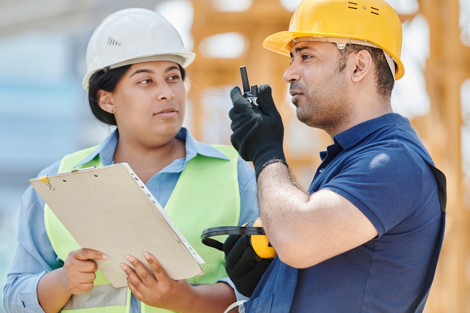 Two site workers in hard hats checking a clipboard and speaking on a radio